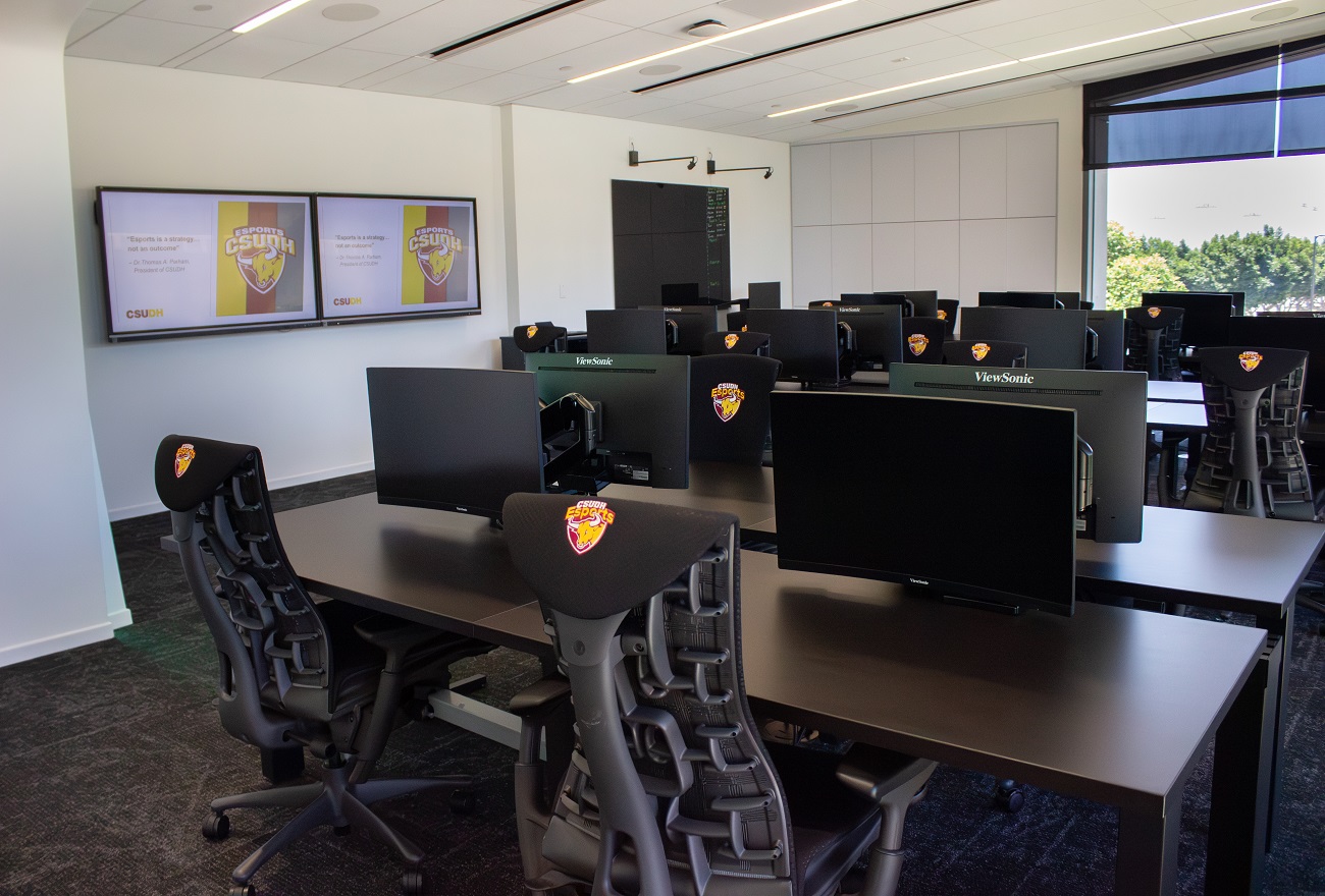 A few of a room with two monitors mounted on the wall displaying the CSUDH Esports logo. In the room there are several tables with monitors on tables with chairs at each table. These chairs have CSUDH Esports branding as well.