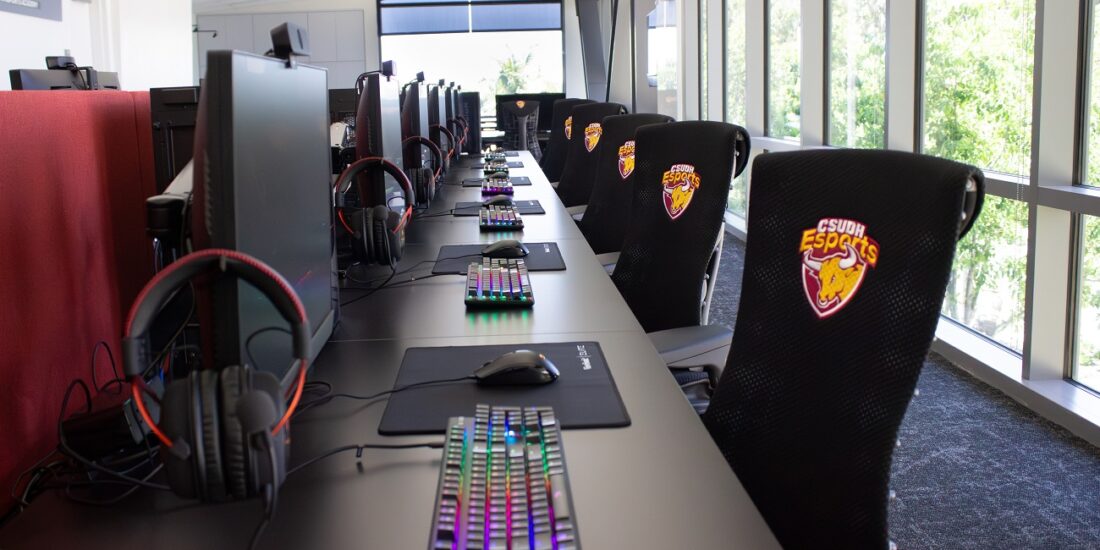 A long table with a row of Monitors. There are headphones hanging next to each monitor. There are designer led keyboards with rainbow colors lit up. There are CSUDH Esports-branded chairs in front of each monitor.