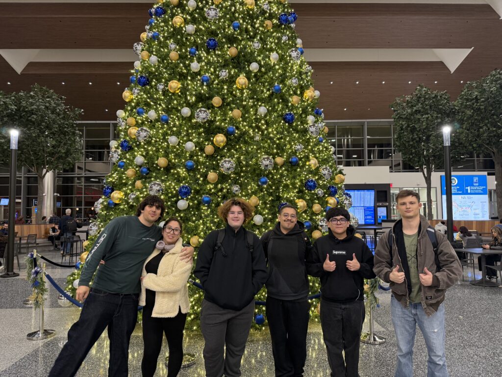 Six students pose together in front of a tall, brightly lit Christmas tree decorated with blue, gold, and silver ornaments inside a campus common area. The group is standing side by side, smiling and giving thumbs-up gestures.