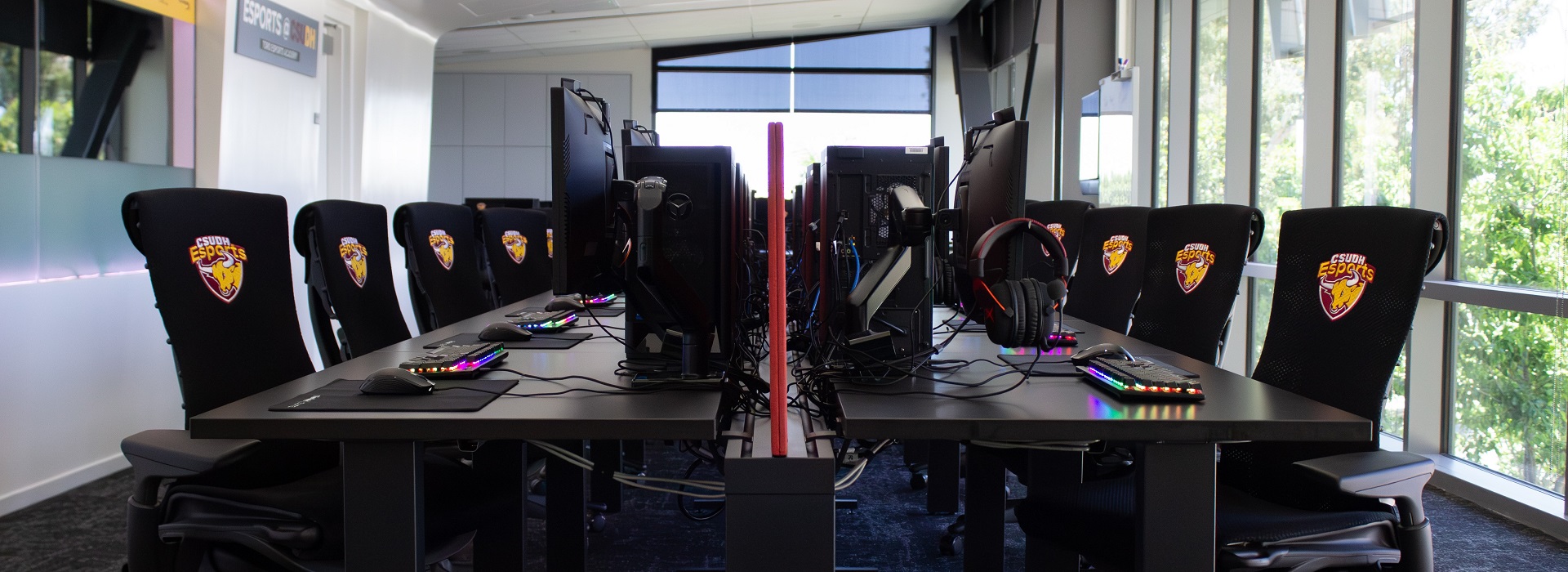 A view of the Toro Esports Academy showing a bank of CPUs with monitors and headphones hanging off to the side. There are chairs witht the CSUDH Esport logo o nthem.