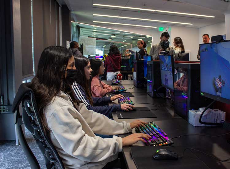 In the foreground, there are three Compton students looking forward at a monitor with their hands on their keyboards. In the background, there is someone taking a picture of the students.