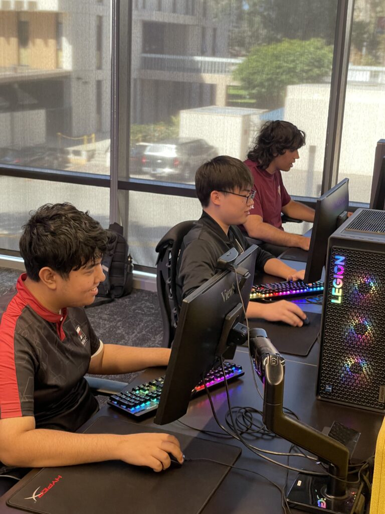 Three students sit at desktop gaming PCs, focused on their screens while using keyboards and mice in a computer lab.