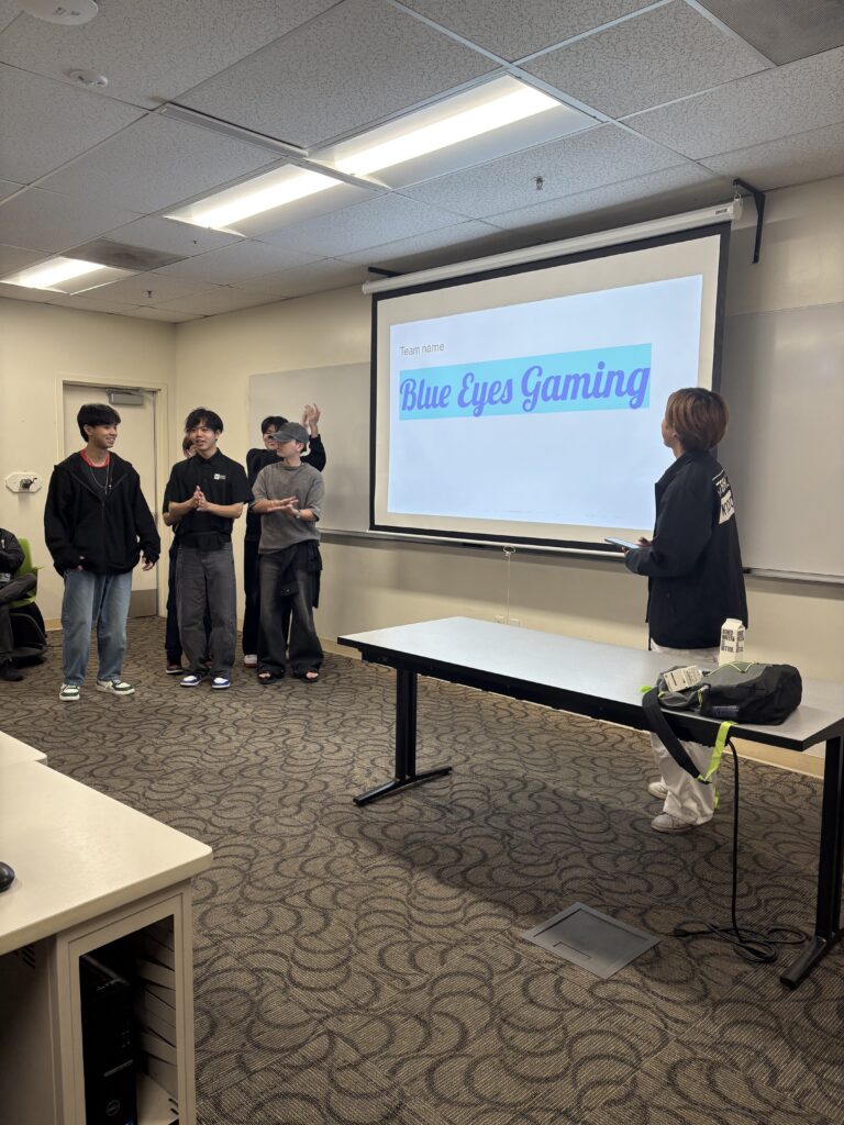 A group of students stands at the front of a classroom presenting a slide titled “Blue Eyes Gaming” while an organizer looks on.
