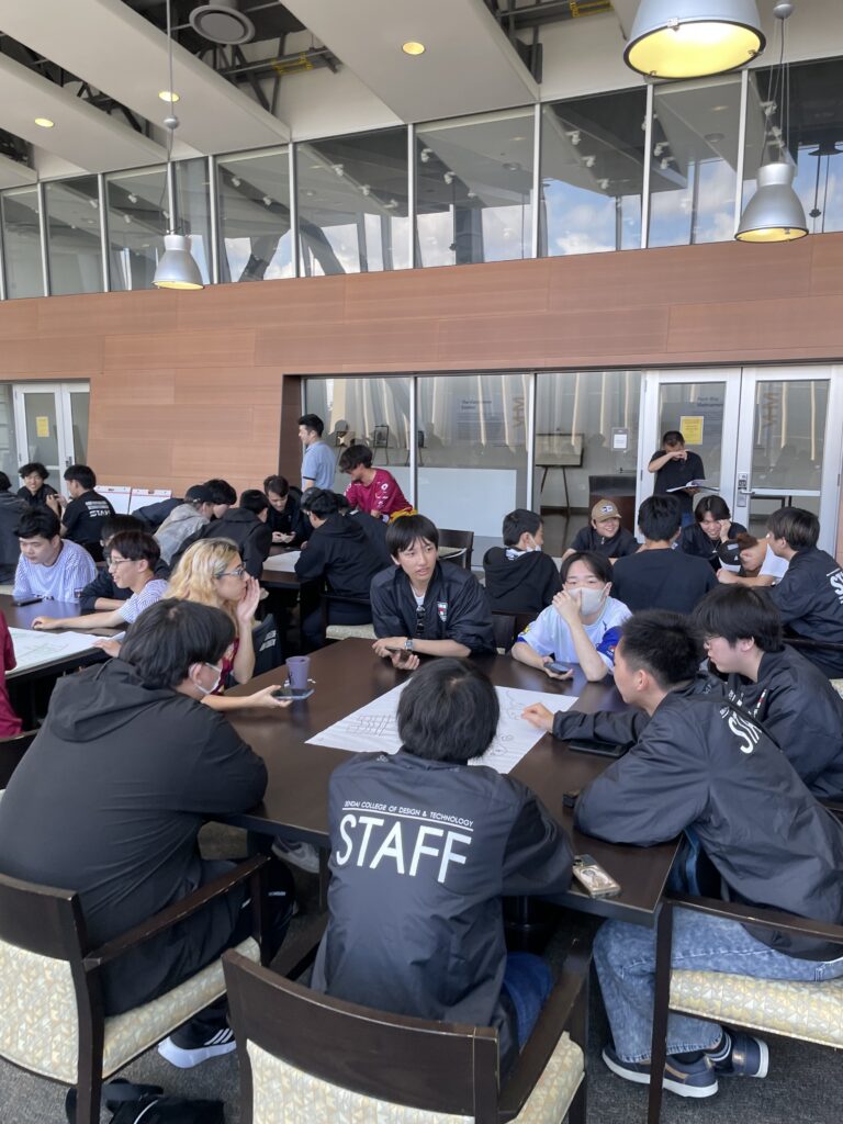 A large group of students wearing matching staff jackets sit around tables collaborating and discussing ideas in a bright, open indoor space.